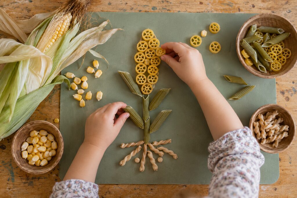 A preschool child building a corn plant with yellow, green and tan pasta loose parts.
